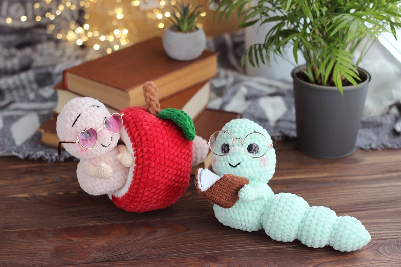 May include: Two crocheted worm plushies, one wearing pink heart-shaped sunglasses and sitting inside a red crocheted apple, the other wearing round glasses and holding a brown crocheted book.