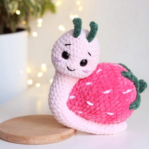 Può includere: Una lumaca rosa all'uncinetto con un gambo verde e un guscio di fragola rosa. La lumaca ha un viso carino con occhi neri e un sorriso.