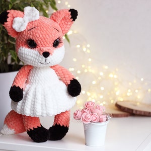 May include: A crocheted fox plush toy wearing a white dress and a white bow. The fox has black eyes, a black nose, and black paws. The toy is sitting on a white surface with a small white bucket of pink flowers in the background.