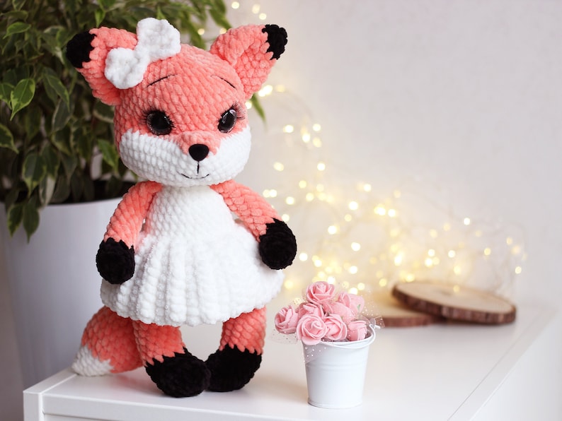 May include: A crocheted fox plush toy wearing a white dress and a white bow. The fox has black eyes, a black nose, and black paws. The toy is sitting on a white surface with a small white bucket of pink flowers in the background.