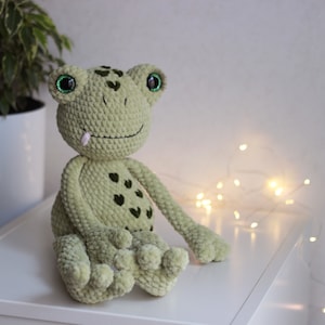May include: A green crocheted frog plush toy with large green eyes and a pink tongue sticking out. The frog is sitting on a white surface with string lights in the background.