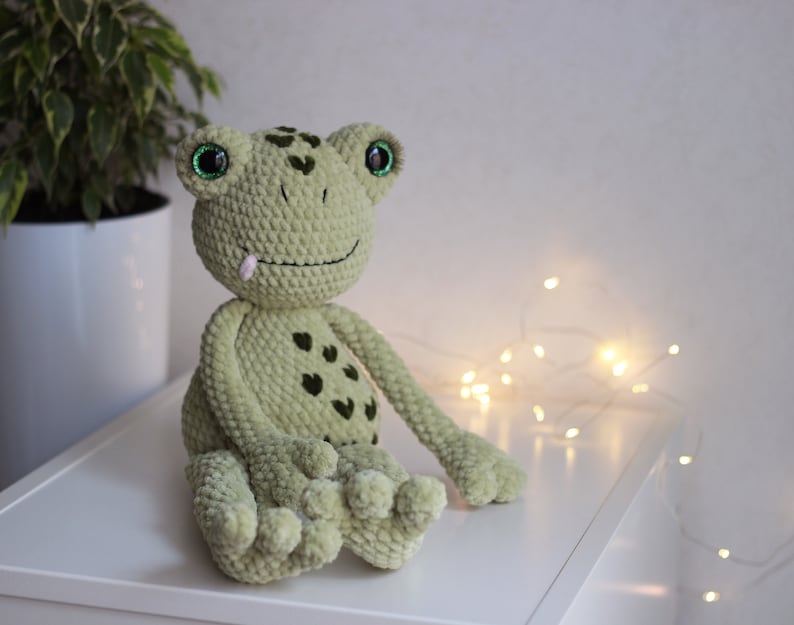 May include: A green crocheted frog plush toy with large green eyes and a pink tongue sticking out. The frog is sitting on a white surface with string lights in the background.