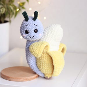 Può includere: Un giocattolo di peluche lavorato a uncinetto a forma di banana con un bruco blu e bianco in cima. Il bruco ha un viso sorridente e antenne verdi.