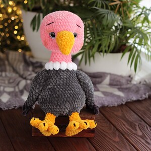May include: A handmade plush vulture toy with a pink head, yellow beak, and black eyes. The vulture has a gray body, white collar, and yellow feet. It is sitting on a wooden surface.