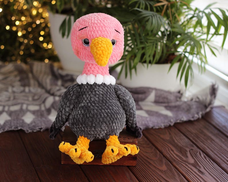 May include: A handmade plush vulture toy with a pink head, yellow beak, and black eyes. The vulture has a gray body, white collar, and yellow feet. It is sitting on a wooden surface.