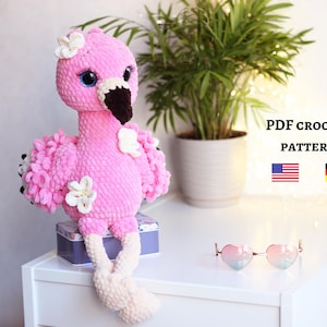 Könnte beinhalten: Rosa gehäkeltes Flamingo-Spielzeug mit weißen Blumen und einem schwarzen Schnabel. Der Flamingo sitzt auf einer weißen Oberfläche mit einer Schachtel und herzförmigen Sonnenbrillen im Hintergrund. Der Text "PDF CROCHET PATTERN" ist in der oberen rechten Ecke des Bildes zu sehen, zusammen mit der amerikanischen und deutschen Flagge.