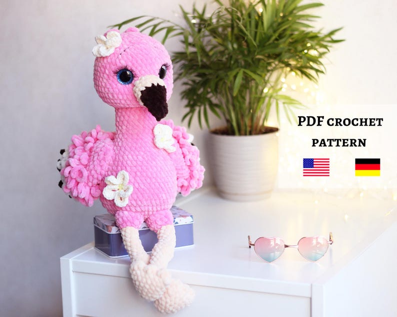 May include: Pink crocheted flamingo toy with white flowers and a black beak. The flamingo is sitting on a white surface with a box and heart-shaped sunglasses in the background. The text "PDF CROCHET PATTERN" is visible in the upper right corner of the image, along with the American and German flags.