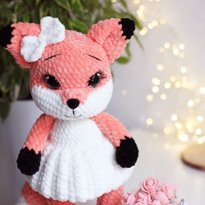May include: A crocheted fox plush toy wearing a white dress and a white bow. The fox has black eyes, a black nose, and black paws.