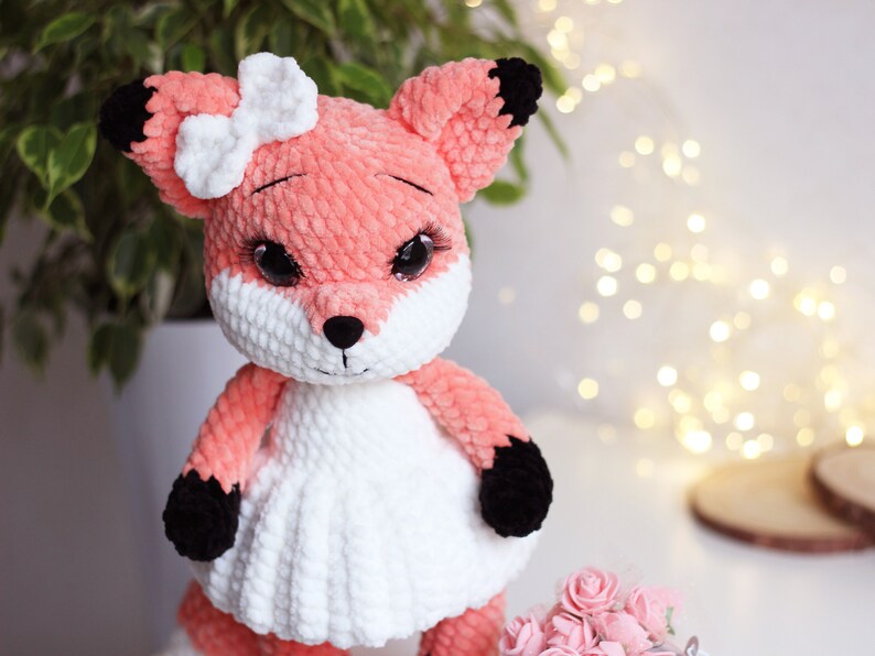 May include: A crocheted fox plush toy wearing a white dress and a white bow. The fox has black eyes, a black nose, and black paws.