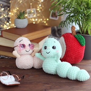 May include: Two crocheted worm plushies, one pink with pink sunglasses and one green, sitting on a wooden surface with a stack of books and a crocheted apple behind them.