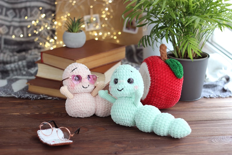 May include: Two crocheted worm plushies, one pink with pink sunglasses and one green, sitting on a wooden surface with a stack of books and a crocheted apple behind them.
