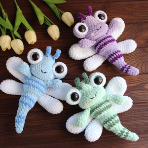 Puede incluir: Tres juguetes de libélula de peluche hechos de hilo suave en azul claro, lavanda y verde menta. Cada juguete presenta grandes ojos de plástico negro, pequeñas antenas y cuatro alas blancas. Los cuerpos tienen rayas de tonos más oscuros.
