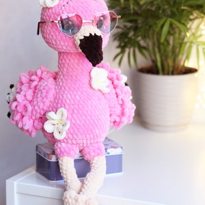 May include: A pink crocheted flamingo toy wearing heart-shaped sunglasses and white flowers. The flamingo is sitting on a white surface with a gray box behind it.