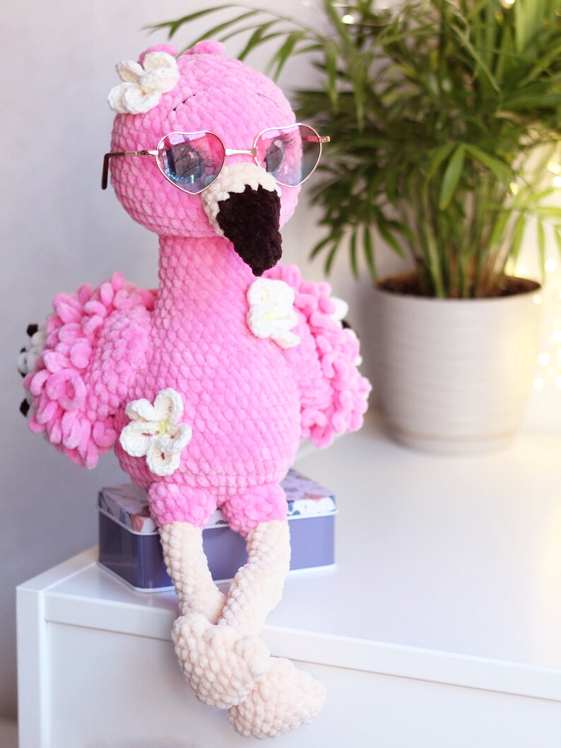 May include: A pink crocheted flamingo toy wearing heart-shaped sunglasses and white flowers. The flamingo is sitting on a white surface with a gray box behind it.