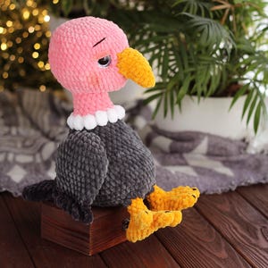 May include: A handmade crochet vulture doll with a pink head, yellow beak and feet, and gray body. The vulture has a white collar and is sitting on a small wooden box. The background includes a blurred string of lights and a houseplant.