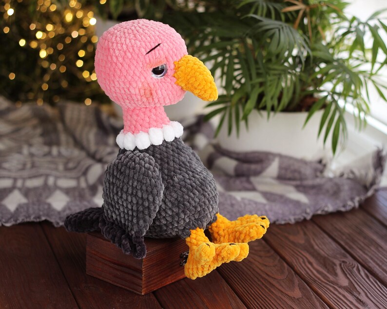 May include: A handmade crochet vulture doll with a pink head, yellow beak and feet, and gray body. The vulture has a white collar and is sitting on a small wooden box. The background includes a blurred string of lights and a houseplant.