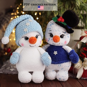May include: Two crocheted snowman dolls. The left snowman wears a light blue hat with white snowflake accents. The right snowman wears a black top hat with holly and a dark blue sweater with a white snowflake. Both have orange noses.