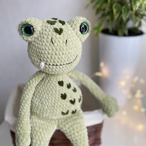 May include: A green crocheted frog plush toy with large green eyes and a pink tongue sticking out. The frog is sitting on a wicker basket.