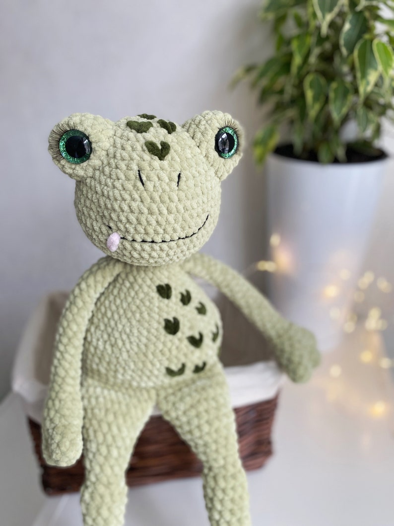 May include: A green crocheted frog plush toy with large green eyes and a pink tongue sticking out. The frog is sitting on a wicker basket.