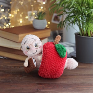 May include: A crocheted worm wearing glasses and holding a book is peeking out of a red crocheted apple.