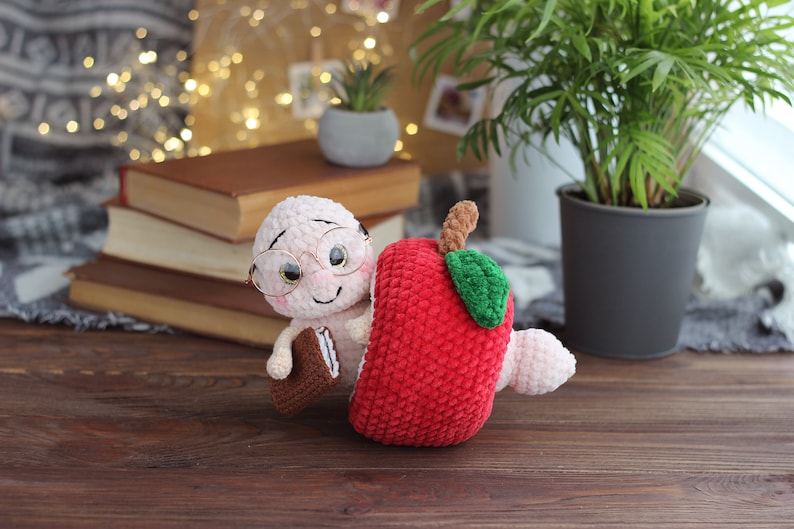 May include: A crocheted worm wearing glasses and holding a book is peeking out of a red crocheted apple.