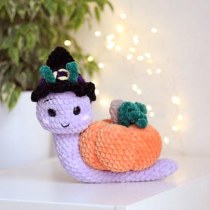 Può includere: Una lumaca di uncinetto viola che indossa un cappello da strega nero con accenti verdi e trasporta una zucca arancione all'uncinetto.