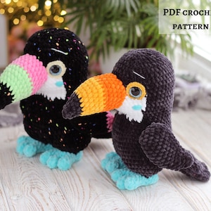 Könnte beinhalten: Zwei gehäkelte Tukan-Plüschtiere. Einer ist schwarz mit bunten Sprenkeln, einem rosa, grünen und schwarzen Schnabel und blauen Füßen. Der andere ist dunkelgrau mit einem orangefarbenen und gelben Schnabel und blauen Füßen. Der Text "PDF CROCHET PATTERN" ist sichtbar.