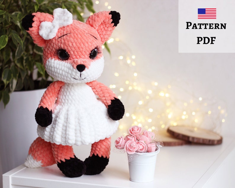 May include: A crocheted fox doll wearing a white dress and a pink bow. The fox has black eyes and a black nose. The doll is sitting on a white surface with a small white pot of pink flowers in the background.