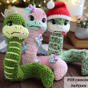 Può includere: Tre serpenti di peluche realizzati in filato morbido. Il primo è verde con una corona dorata, il secondo è rosa con accenti floreali blu e viola, il terzo è verde e bianco screziato con un cappello da Babbo Natale rosso e bianco. Testo: PDF CROCHET PATTERN.