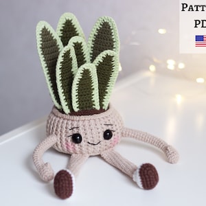 May include: Crocheted amigurumi toy of a potted plant with a smiling face and brown legs. The plant has green and brown leaves and is in a brown pot. The toy is sitting on a white surface.