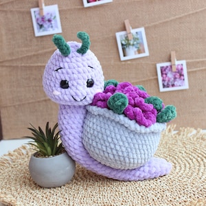 以下が含まれることがあります： 緑の葉のついた紫の花の入ったバスケットを運ぶ、紫色の毛糸で編まれたカタツムリ。カタツムリは黒い目をした笑顔で、織り込まれたマットの上に座っています。