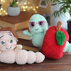 May include: Three crocheted stuffed toys: a pink worm wearing glasses and a brown book on its head, a green worm wearing sunglasses, and a red apple with a green leaf.