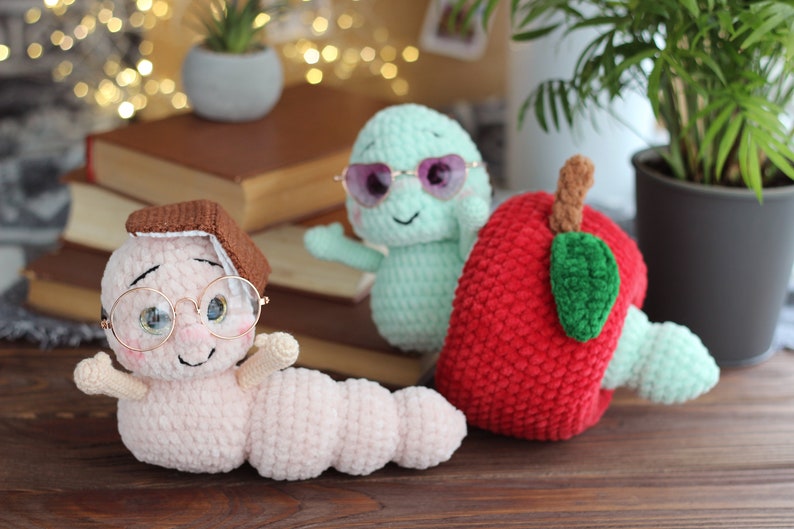 May include: Three crocheted stuffed toys: a pink worm wearing glasses and a brown book on its head, a green worm wearing sunglasses, and a red apple with a green leaf.