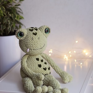 May include: A green crocheted frog toy with large green eyes and a pink tongue sticking out. The frog is sitting on a white surface.