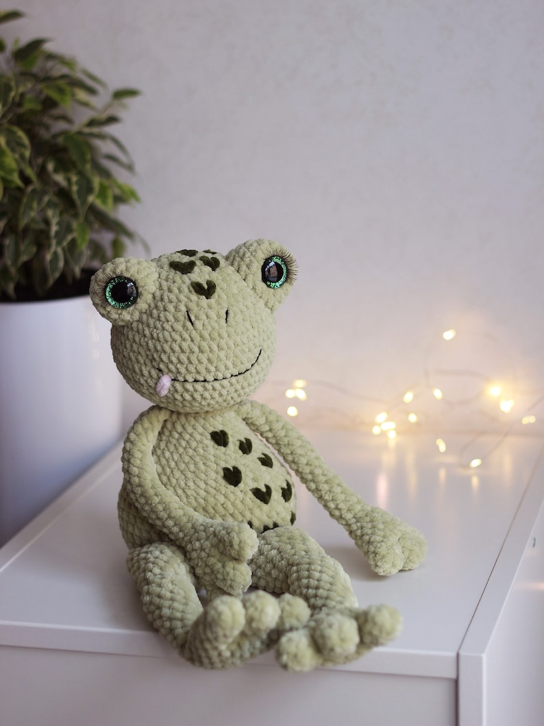 May include: A green crocheted frog toy with large green eyes and a pink tongue sticking out. The frog is sitting on a white surface.