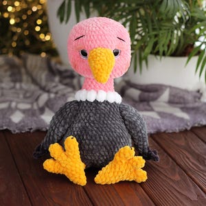 May include: A handmade plush vulture toy. The vulture has a pink head, a yellow beak, and a gray body. It has yellow feet and a white collar. The toy is sitting on a wooden surface.