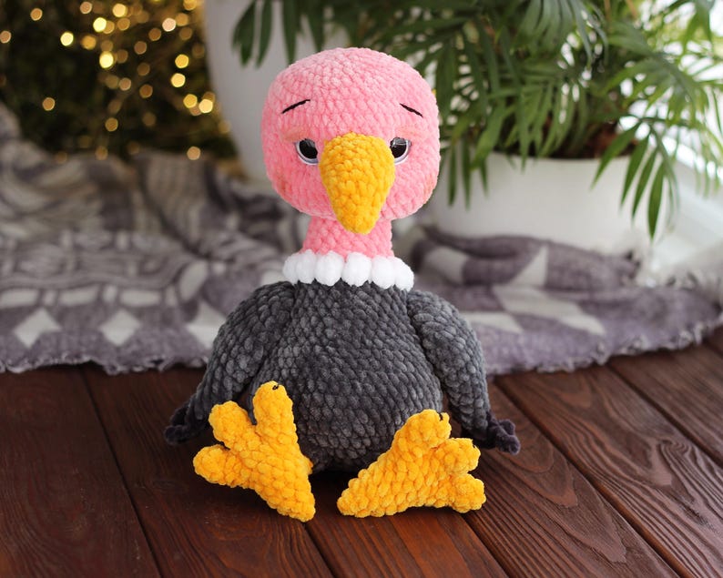 May include: A handmade plush vulture toy. The vulture has a pink head, a yellow beak, and a gray body. It has yellow feet and a white collar. The toy is sitting on a wooden surface.
