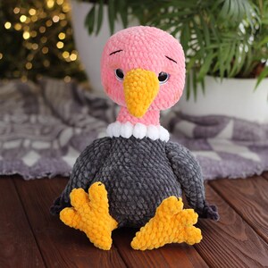 May include: A handmade plush vulture toy with a pink head, yellow beak, and gray body. The vulture has a white collar and yellow feet. The toy is sitting on a wooden surface.