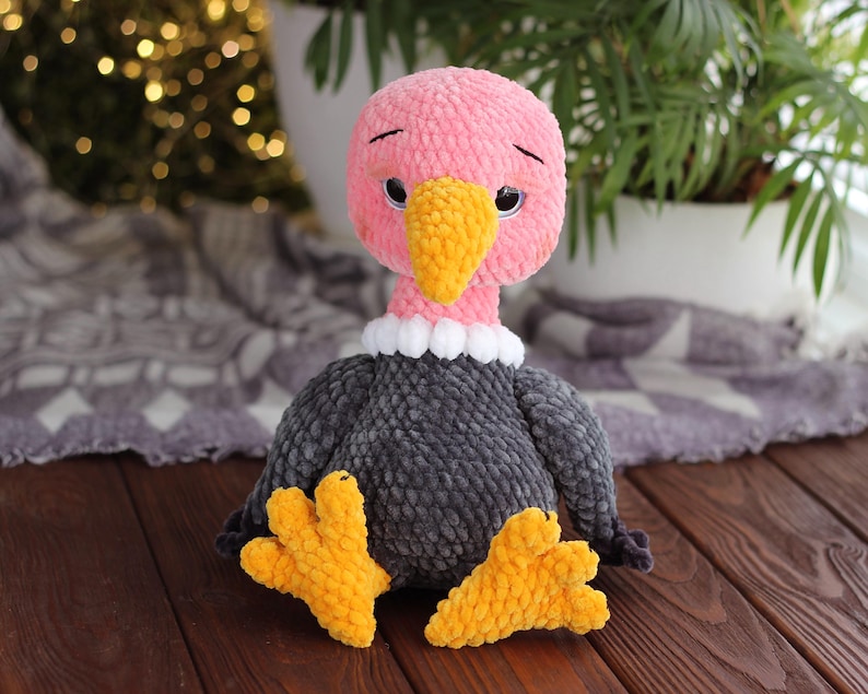 May include: A handmade plush vulture toy with a pink head, yellow beak, and gray body. The vulture has a white collar and yellow feet. The toy is sitting on a wooden surface.