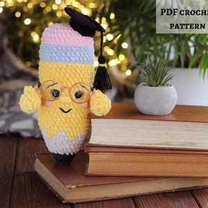 May include: A handmade crochet pencil doll wearing glasses and a graduation cap, with a smiling face. The pencil is yellow, pink, and blue, and is sitting on top of a stack of old books. The text "PDF CROCHET PATTERN" is visible in the image.