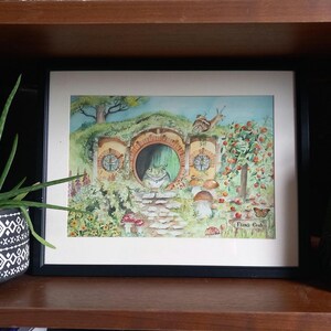 May include: A framed watercolour painting depicting a frog's home with a green door and a frog looking out. The artwork includes a stone path, mushrooms, and a snail. The words "Frog End" are written in the lower right corner.