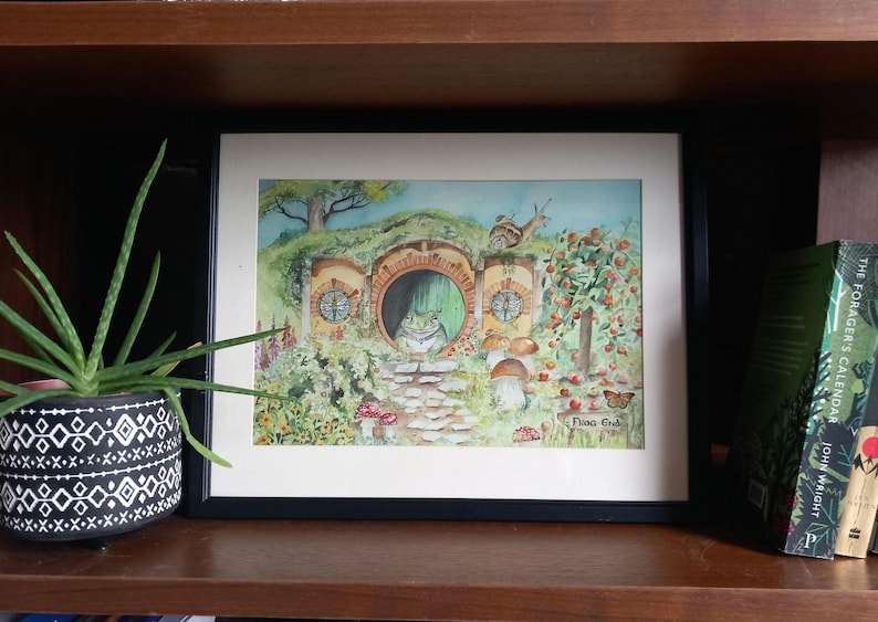May include: A framed watercolour painting depicting a frog's home with a green door and a frog looking out. The artwork includes a stone path, mushrooms, and a snail. The words "Frog End" are written in the lower right corner.