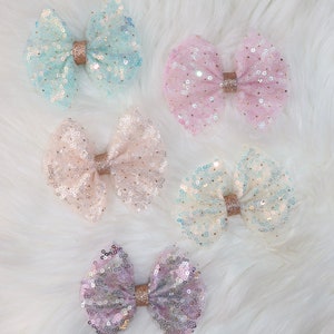 May include: Four sparkly hair bows in different colors: light blue, pink, beige, and purple. Each bow is made of tulle and sequins and has a gold ribbon in the center.