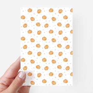 May include: A white card featuring a repeating pattern of orange pumpkins and small white crescent moons. The pumpkins have green stems and are spread across the card. The card is held by a hand.