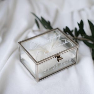 Puede incluir: Una caja de anillos de vidrio transparente con marco de metal plateado, sobre tela blanca. La caja está grabada con nombres y la fecha 22.08.2026. Una almohada blanca está dentro.