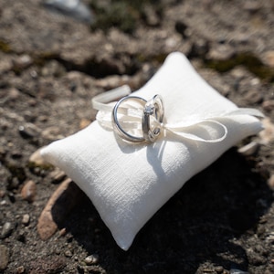 Könnte beinhalten: Zwei goldene Eheringe mit einem Diamanten auf einem weißen Kissen. Das Kissen liegt auf einem Bett aus kleinen Steinen.