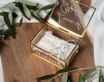 May include: A rectangular glass box with a gold metal frame, featuring a white satin cushion inside. The lid has white script text, and the front glass panel displays the date 25.10.2025 in white. A small sprig of white flowers rests on the cushion.