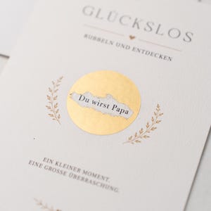 Peut inclure: Une carte couleur crème avec le texte GLÜCKSLOS, RUBBELN UND ENTDECKEN, et EIN KLEINER MOMENT. EINE GROSSE ÜBERRASCHUNG. Un autocollant à gratter circulaire doré est partiellement retiré, révélant le texte Du wirst Papa en dessous.