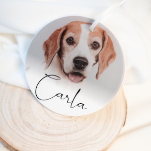 Op de afbeelding: Een ronde, witte ornament met een hondenportret met bruine en witte vacht. De naam "Carla" is in cursief geschreven. Een wit lint is bevestigd om op te hangen. Het ornament rust op een houten schijf.