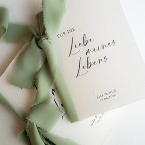 Könnte beinhalten: Cremefarbene Hochzeitseinladungskarten mit zerrissenen Kanten, gebunden mit einem salbeigrünen Band. Die Karten haben eine elegante Kalligraphie mit der Aufschrift "Für die Liebe meines Lebens" und Namen mit Datum.